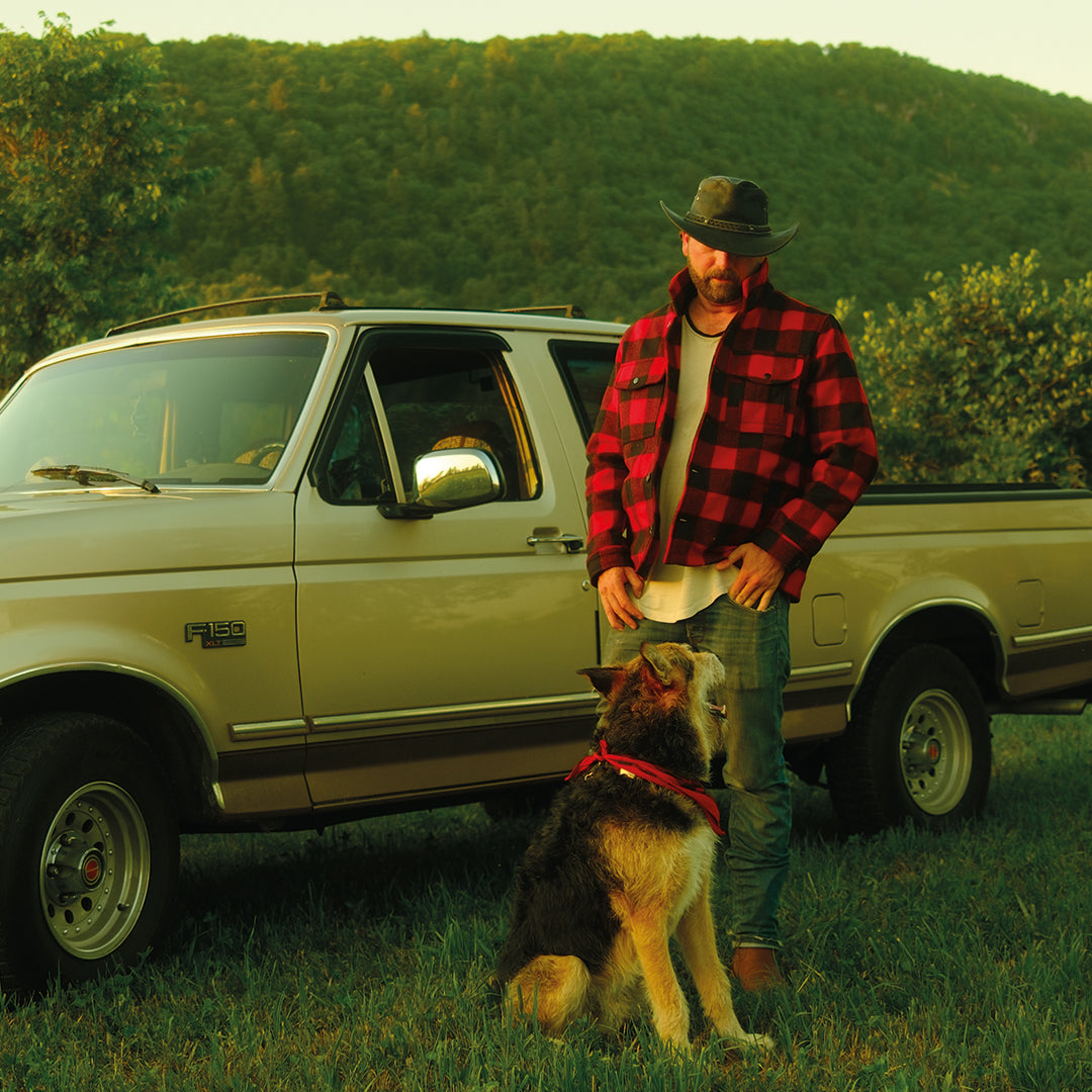 Man in a red plaid shirt and cowboy hat standing next to a beige truck with a dog in a field.
