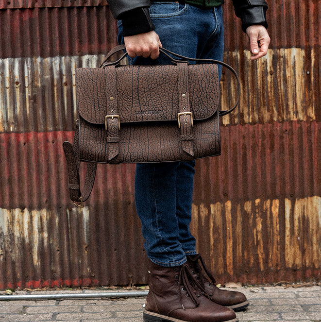 A person wearing a black jacket and blue jeans is holding a brown rust-colored leather messenger bag with brass buckles and a zipper closure.