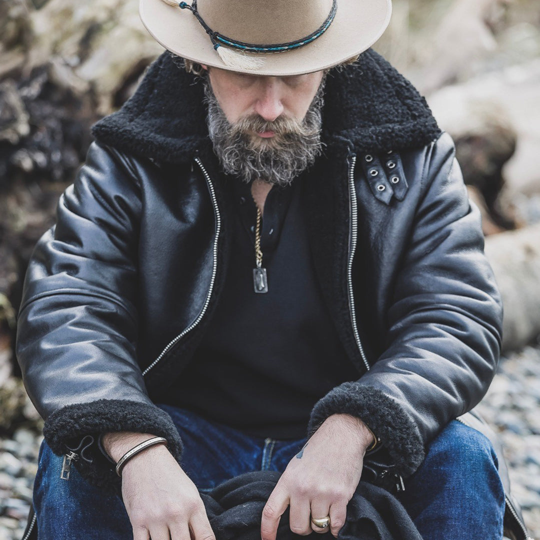 Man wearing a black leather jacket with fur lining, sitting on rocks. Lastwolf B3 Leather Jacket