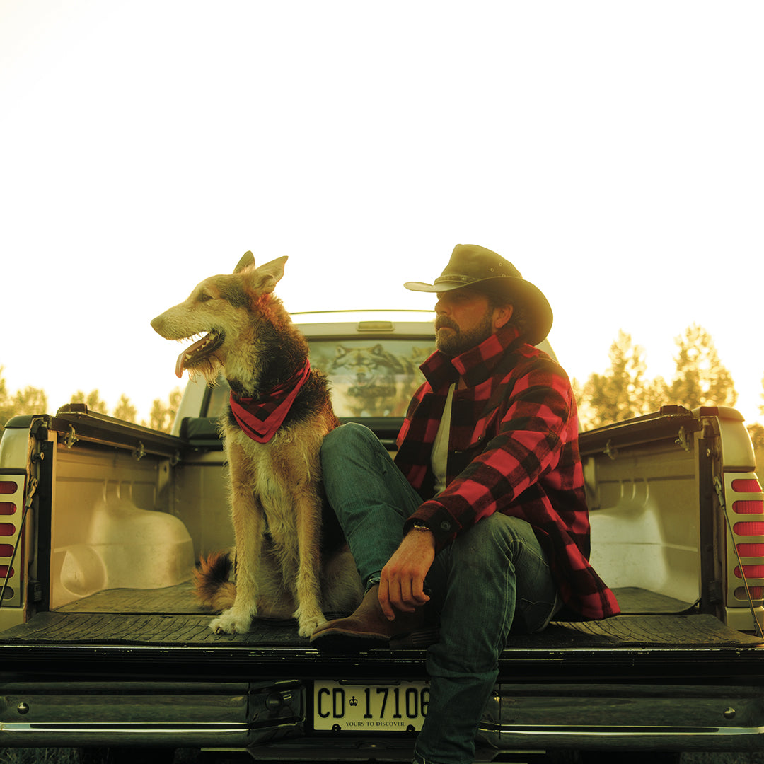 Man in a cowboy hat and plaid jacket sitting in the back of a pickup truck with a dog.