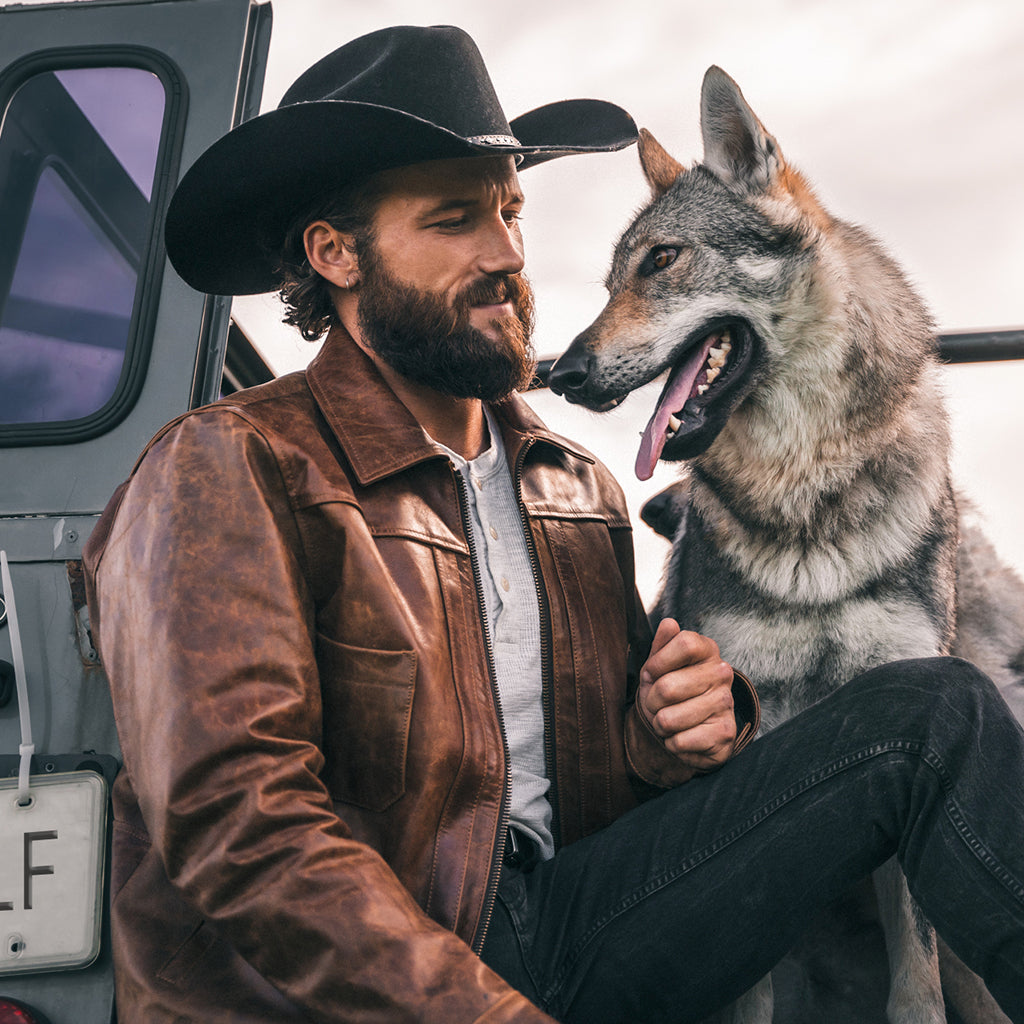 Man in a brown leather jacket beside a gray wolf outdoors