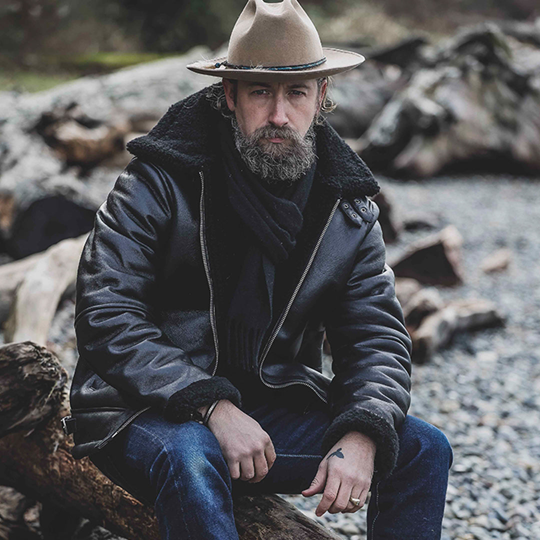 A man sitting outdoors wearing a black shearling leather jacket with a fur hood, paired with a hat and jeans.